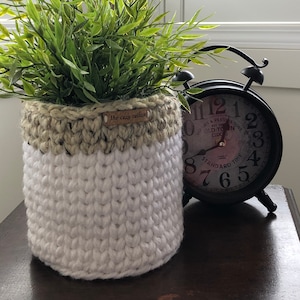 May include: A white crocheted planter with a brown crocheted top, holding a green artificial plant. The planter has a brown label that reads "The cozy cottage". A black alarm clock with white numbers and a white face is sitting on a wooden surface.
