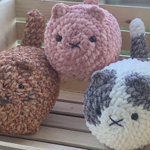 Könnte beinhalten: Drei gehäkelte Katzenplüschtiere in einer Holzkiste. Die Katzenplüschtiere sind braun, rosa und weiß mit grauen Flecken. Jedes Plüschtier hat eine schwarze gestickte Nase und Augen.