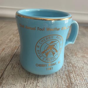 May include: A light blue ceramic mug with a gold rim and handle. The mug features gold text that reads "3rd Annual Foul Weather Rifle Co." and "N. Cherry Point Rifle & Pistol Club, Cherry Point, NC 1981."