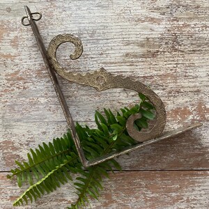 May include: An antique-style, metal wall shelf with a decorative scroll design and a small shelf. The shelf is a muted gold color and is adorned with green fern leaves. The shelf is mounted on a wooden surface.