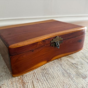 May include: A rectangular wooden box featuring a rich reddish-brown grain pattern with lighter amber accents. The lid is secured by a central antique brass-toned metal latch. The box has smooth, rounded edges and a polished finish throughout.