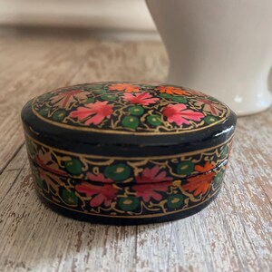 May include: A small, oval-shaped trinket box with a black base and a colorful floral design. The box is decorated with pink, orange, green, and gold paint.