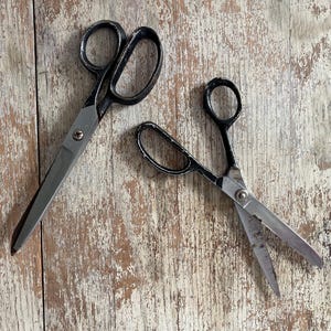 May include: Two pairs of vintage scissors with black handles and silver blades rest on a distressed wooden surface. One pair is larger, while the other is smaller and open. The scissors are likely used for fabric or paper.