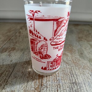 May include: A vintage white glass with a red graphic design featuring landmarks and text. The design includes illustrations of a Native American, a building, and the text "THE TAR HEEL STATE". The glass is on a wooden surface.