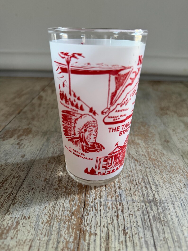 May include: A vintage white glass with a red graphic design featuring landmarks and text. The design includes illustrations of a Native American, a building, and the text "THE TAR HEEL STATE". The glass is on a wooden surface.