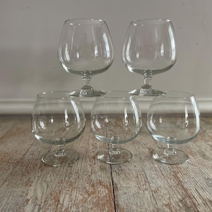 May include: Five clear, stemmed brandy snifters arranged on a light-colored wooden surface. Two glasses are stacked on top of three others. The glasses have a rounded bowl shape and a short stem.