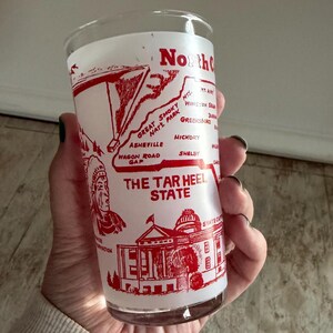 May include: A vintage glass featuring a red and white design with the text "North Carolina" and "The Tar Heel State." The design includes a map, an illustration of a Native American, and a state capitol building.