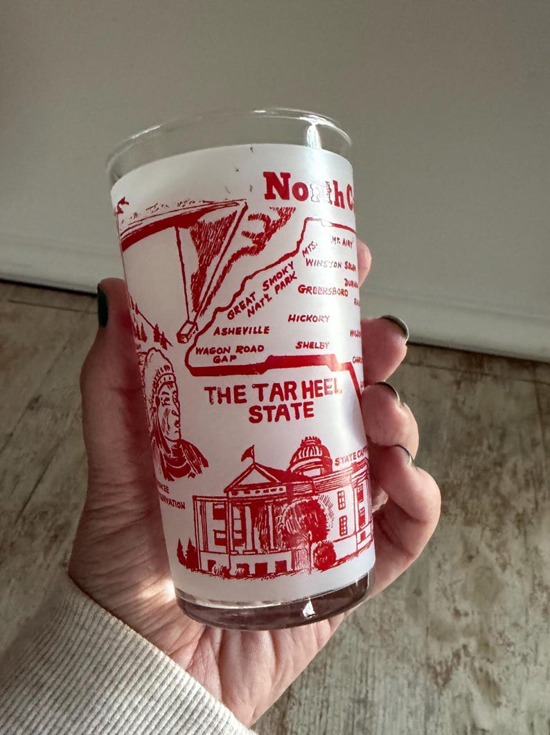 May include: A vintage glass featuring a red and white design with the text "North Carolina" and "The Tar Heel State." The design includes a map, an illustration of a Native American, and a state capitol building.