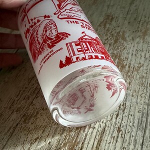 May include: A clear glass tumbler with a white background and red graphics. The graphics include a Native American figure, a building, and text that reads "The Tar Heel State" and "Cherokee Indian Reservation."
