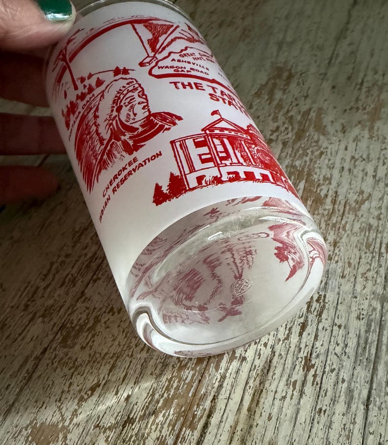 May include: A clear glass tumbler with a white background and red graphics. The graphics include a Native American figure, a building, and text that reads "The Tar Heel State" and "Cherokee Indian Reservation."