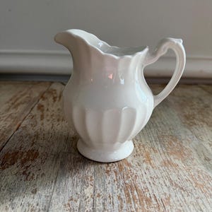 May include: A white ceramic creamer pitcher with a scalloped rim and a curved handle. The pitcher has vertical ribbed detailing and sits on a small, round base. The background is a light-colored, distressed wooden surface.