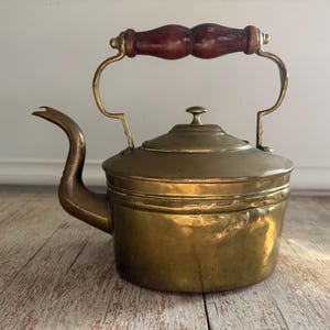 May include: Antique brass kettle with a curved spout and a wooden handle. The kettle has a round body with a lid and a handle that arches over the top. The brass has a warm, golden hue, and the wood handle is a dark brown.