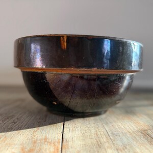 May include: A dark brown, glazed ceramic bowl with a wide rim and a rounded base. The bowl has a rustic, antique appearance, with visible imperfections and variations in the glaze. The bowl sits on a wooden surface.
