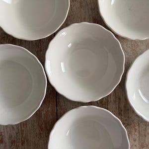 May include: A set of six white ceramic bowls arranged in a circular pattern. Each bowl features a smooth, glossy finish and a distinct scalloped edge design. The bowls are uniform in size and color, presenting a clean, simple aesthetic for dining.