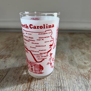 May include: A clear glass with a white interior and a red design featuring the words "North Carolina" and the names of various cities. The design includes a map outline of the state and a depiction of the State Capitol.