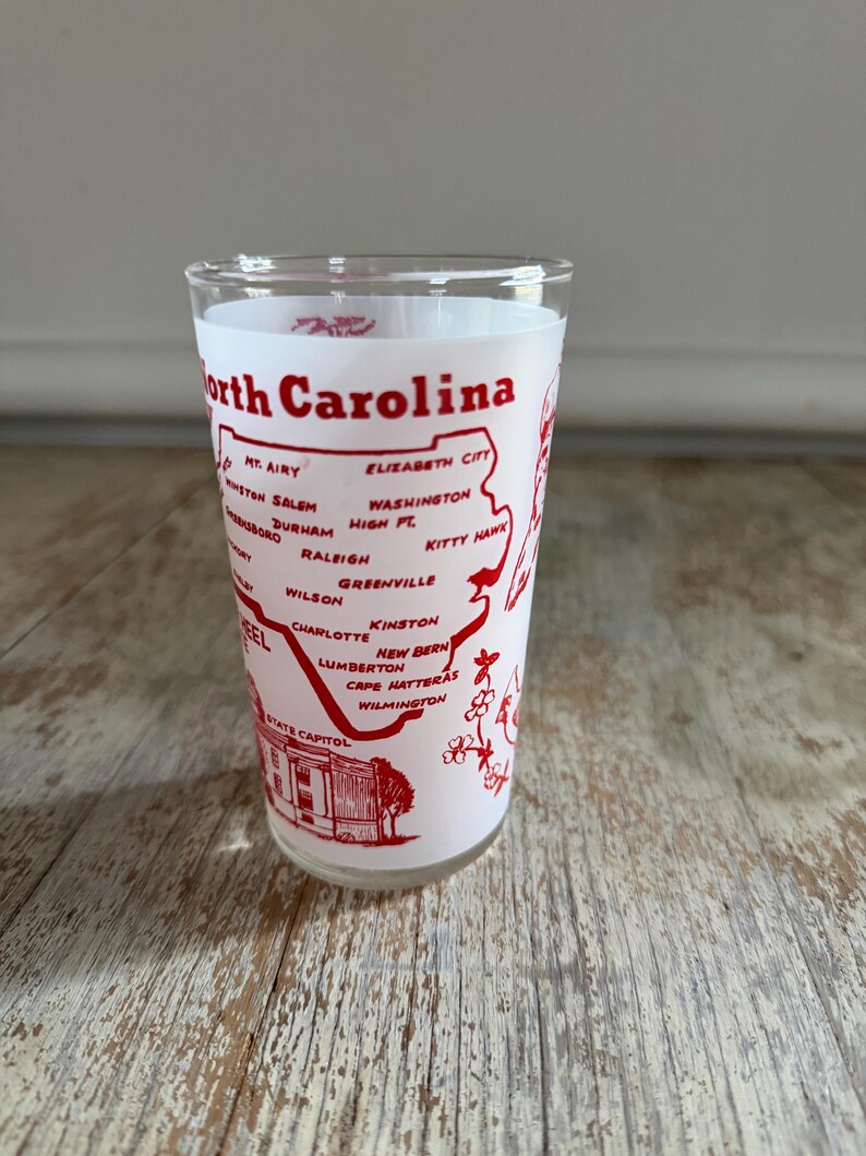 May include: A clear glass with a white interior and a red design featuring the words "North Carolina" and the names of various cities. The design includes a map outline of the state and a depiction of the State Capitol.