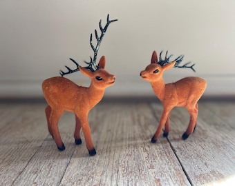 May include: Two orange-brown deer figurines with black antlers and black hooves stand on a light-colored wooden surface. The deer are facing each other, creating a charming decorative display. The figurines have a soft, velvety texture.