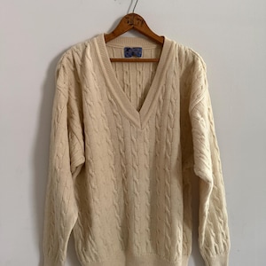 May include: Cream-colored cable knit sweater with a deep V-neck. The sweater is hanging on a wooden hanger. The knit pattern is visible across the front and sleeves. A small label is visible at the back of the neck.