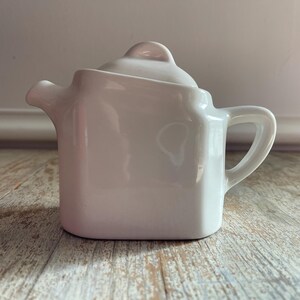 May include: A white ceramic teapot with a square base and a curved handle. The teapot has a lid with a small knob and a spout for pouring. The teapot is sitting on a wooden surface.