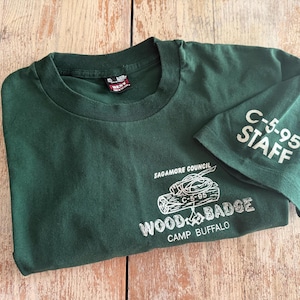 May include: A dark green short-sleeve t-shirt featuring white screen-printed graphics. The front displays the text Sagamore Council, Wood Badge, and Camp Buffalo with a log illustration. The sleeve is printed with the text C-5-95 STAFF.