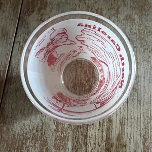 May include: Clear glass bowl with red text and illustrations. The bowl has a wide rim and a circular opening at the bottom. The text appears to be a list of ingredients or instructions. The bowl is sitting on a wooden surface.