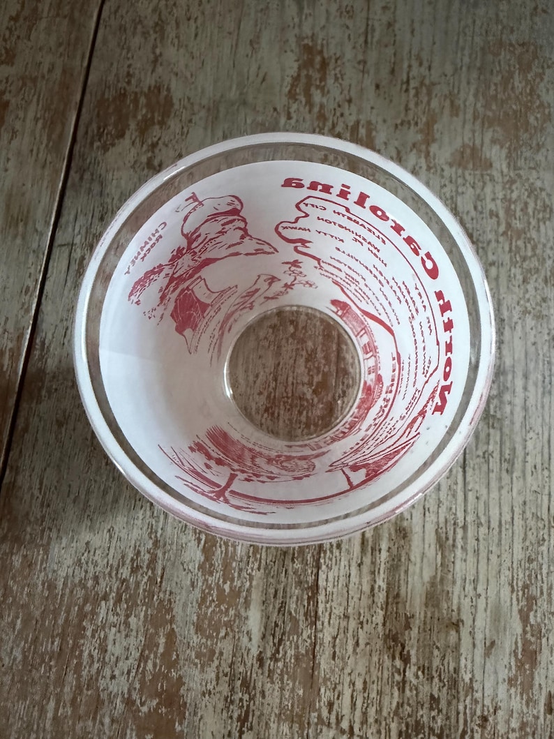 May include: Clear glass bowl with red text and illustrations. The bowl has a wide rim and a circular opening at the bottom. The text appears to be a list of ingredients or instructions. The bowl is sitting on a wooden surface.