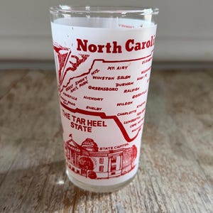 May include: A clear glass with a red design featuring the words "North Carolina" and "The Tar Heel State." The design includes a map of North Carolina with city names and a depiction of the State Capitol building.