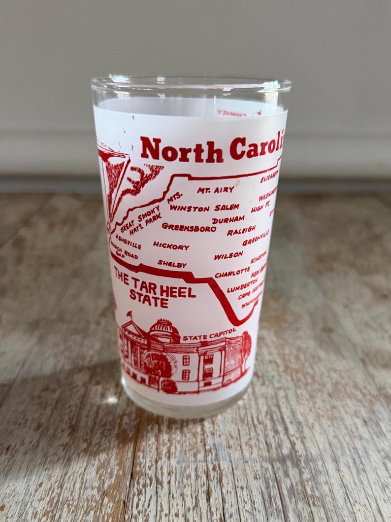 May include: A clear glass with a red design featuring the words "North Carolina" and "The Tar Heel State." The design includes a map of North Carolina with city names and a depiction of the State Capitol building.