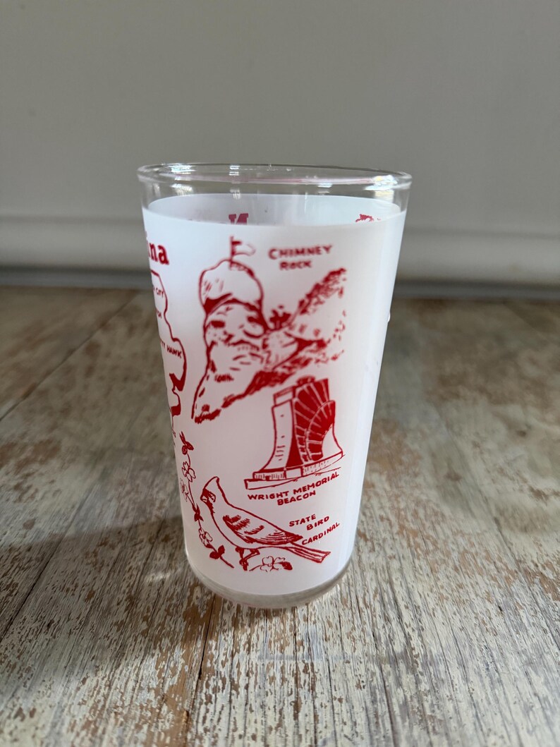 May include: A clear glass tumbler with a white background and red illustrations. The tumbler features images of Chimney Rock, Wright Memorial Beacon, and a cardinal. The text "na" is visible on the side.