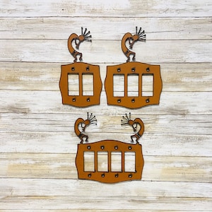 May include: Three decorative metal light switch covers with a Kokopelli design. The covers are a warm brown color and feature cut-out rectangles for the switches. Each cover has a Kokopelli figure at the top.