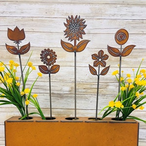 May include: Five metal flower stems in a rusty brown planter. The flowers are different shapes and sizes, including a sunflower, a tulip, and a daisy. The planter has five holes for the stems.