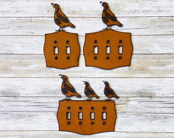 May include: Three sets of light brown wooden light switch covers with a quail bird design. Each cover has a different number of switches. The covers are on a white wood background.