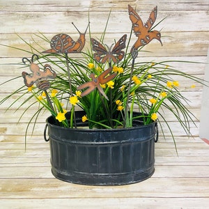 May include: A black metal planter with a rustic finish, filled with green plants and yellow flowers. Five metal garden stakes with various animal and insect designs are placed in the planter. The designs include a snail, a butterfly, a dragonfly, a hummingbird, and a bee.