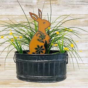 May include: A black metal planter with a rustic finish and two handles. A cutout metal bunny with floral details is placed inside the planter. The planter is filled with green grass and yellow flowers.