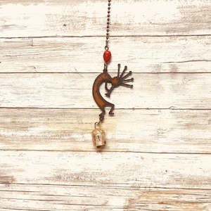 May include: A metal Kokopelli wind chime with a red bead and a gold bell hanging from a chain against a white wood background.