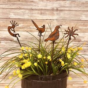 May include: Four metal garden stakes with a rustic finish. The stakes feature a stylized sun, a quail, a roadrunner, and a Native American dancer. The stakes are displayed in a black metal planter with green grass and yellow flowers.