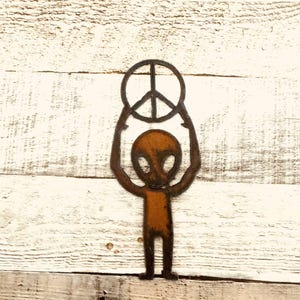 May include: A rusty metal alien figure holding a peace sign above its head. The alien has large oval eyes and a small body. The peace sign is a circle with a three-pronged symbol inside. The background is a white wooden surface.