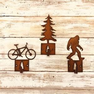 May include: Three metal hooks in the shape of a pine tree, a bicycle, and a Bigfoot. The hooks are mounted on a white wooden wall.