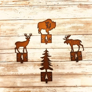 May include: Four rustic metal hooks in the shape of a deer, moose, buffalo, and pine tree. The hooks are mounted on a white wooden background.