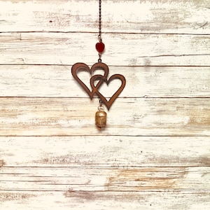 May include: A rustic wooden heart-shaped wind chime with a bell hanging from the bottom. The chime is made of metal and has a red heart charm at the top. The background is a whitewashed wooden wall.