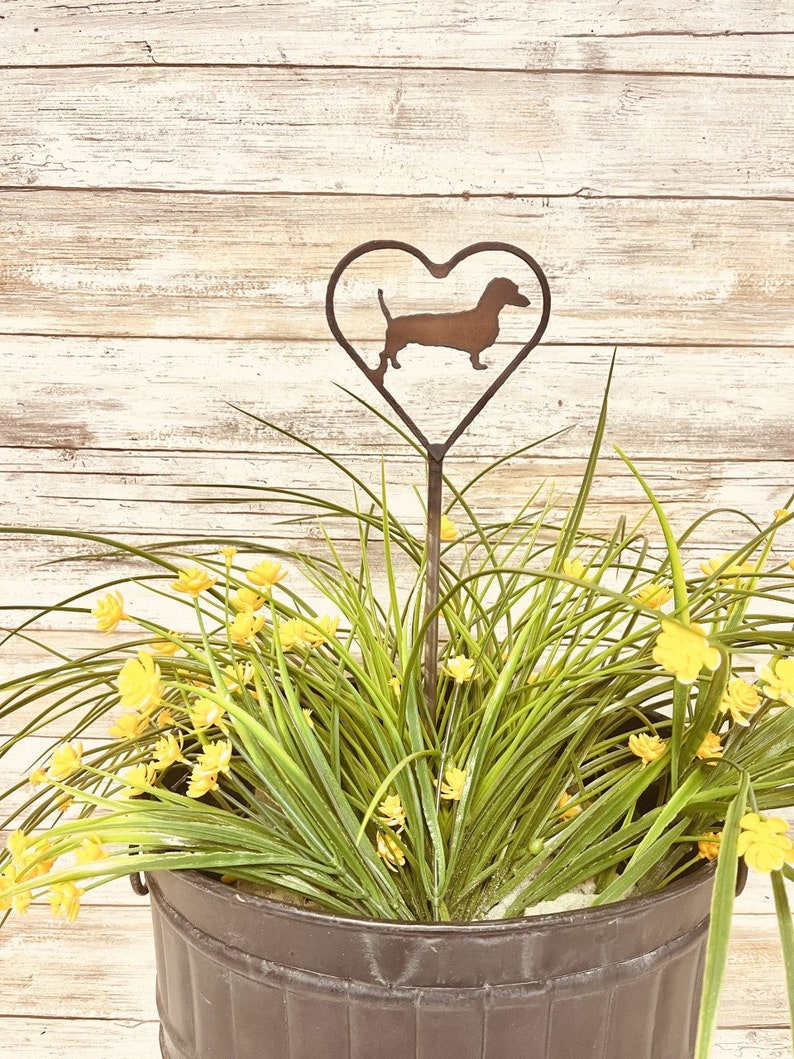 K&ouml;nnte beinhalten: Ein Metall-Gartenpfahl mit einer herzf&ouml;rmigen Silhouette eines Dackelhundes. Der Pfahl steht in einem Topf mit gr&uuml;nen Pflanzen und gelben Bl&uuml;ten.