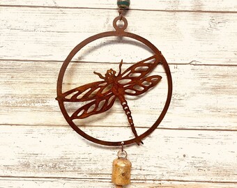 May include: A rusty metal dragonfly wind chime with a bell hanging from the bottom. The dragonfly is in a circle and the chime is hanging from a chain with a green bead.