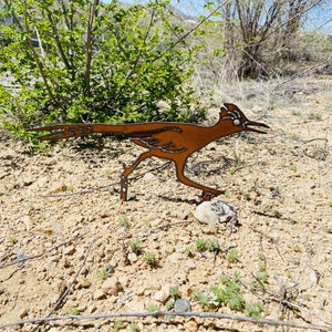 May include: A rusty brown metal sculpture of a roadrunner bird in a running pose. The sculpture is on a sandy ground with some green plants in the background.