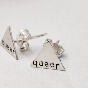 May include: A pair of silver triangle stud earrings with the word "queer" inscribed on each earring.
