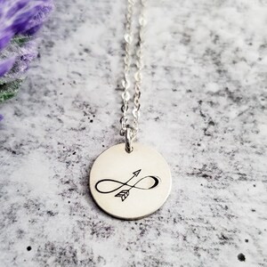 May include: A silver necklace with a round pendant featuring an infinity symbol with an arrow through it.