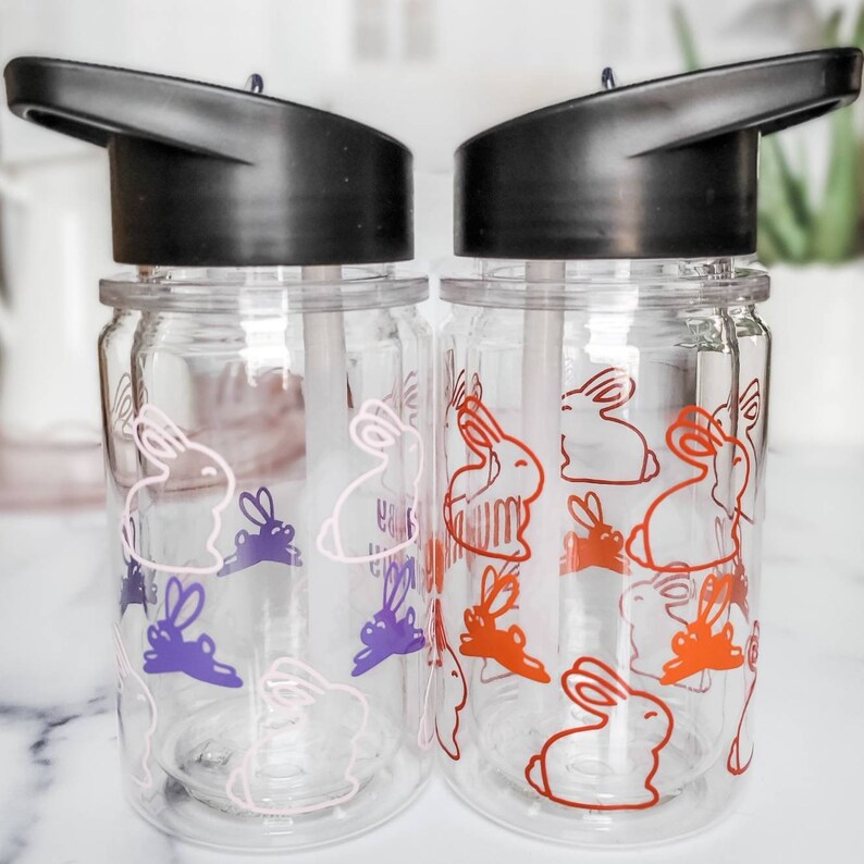 May include: Two clear plastic water bottles with black lids and straws. The bottles have a repeating pattern of pink and purple bunnies on the left bottle and orange bunnies on the right bottle.