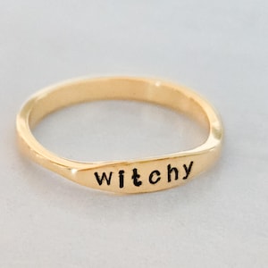 May include: A gold-colored ring with the word "witchy" in black lettering. The ring has a smooth, polished finish and a simple, elegant design. The ring is a piece of jewelry.