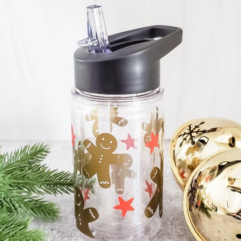 May include: A clear plastic water bottle with a black lid and a straw. The bottle has a gingerbread man design with red stars and is perfect for kids.