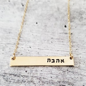 May include: A gold bar necklace with the Hebrew word "אהבה" (love) engraved on it.