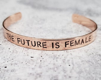 May include: A rose gold colored metal bracelet with the words "THE FUTURE IS FEMALE" engraved on it.